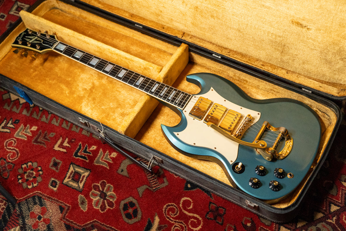 Pelham blue SG Custom in its open case, showing the full body with three gold humbuckers, Bigsby, and white pickguard.