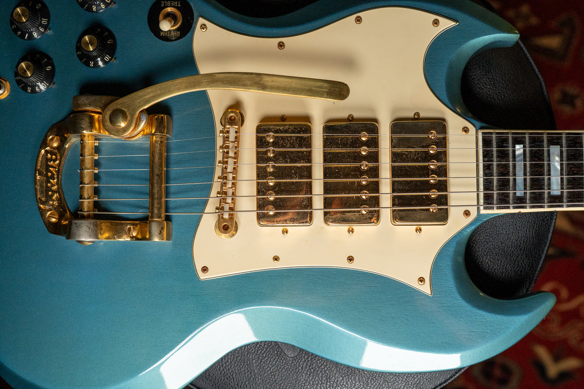 Close-up of a Pelham Blue SG with a cream pickguard, gold Bigsby, three gold humbuckers, and four black control knobs.