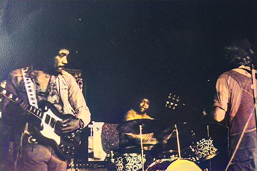 Jimi Hendrix playing a white Gibson SG Custom on stage, with a drummer and another guitarist visible in the background.
