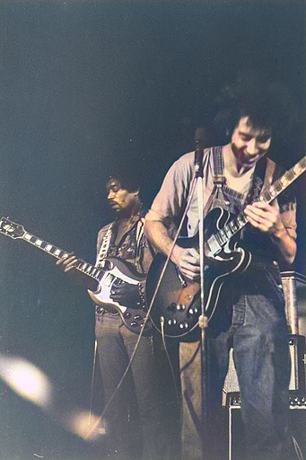 Two men on stage playing electric guitars, one in overalls smiling at his instrument, the other in the background.