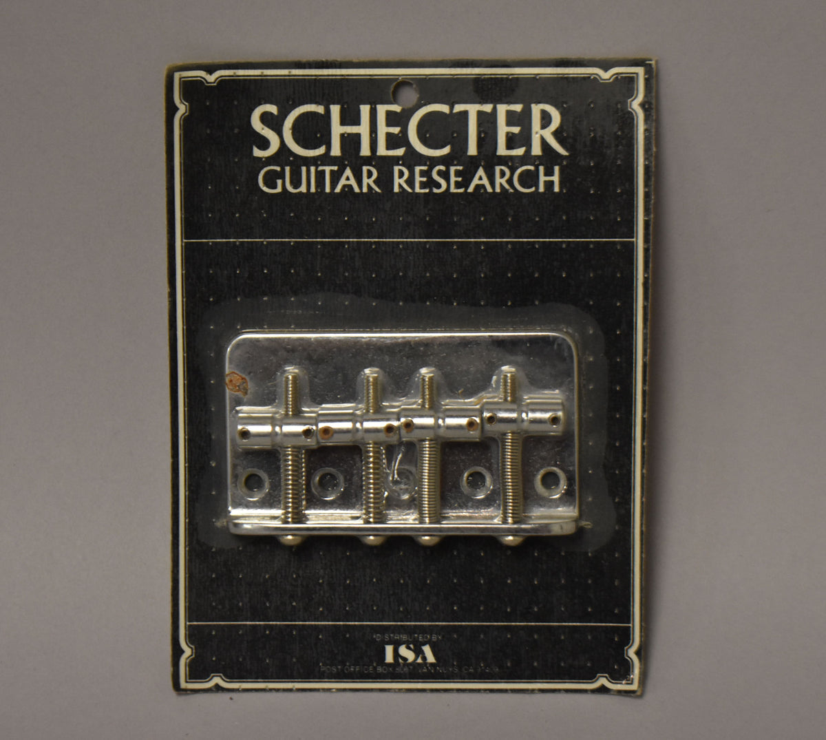 A chrome four-saddle bass bridge in blister packaging, with visible rust spots on the top left of the bridge plate.