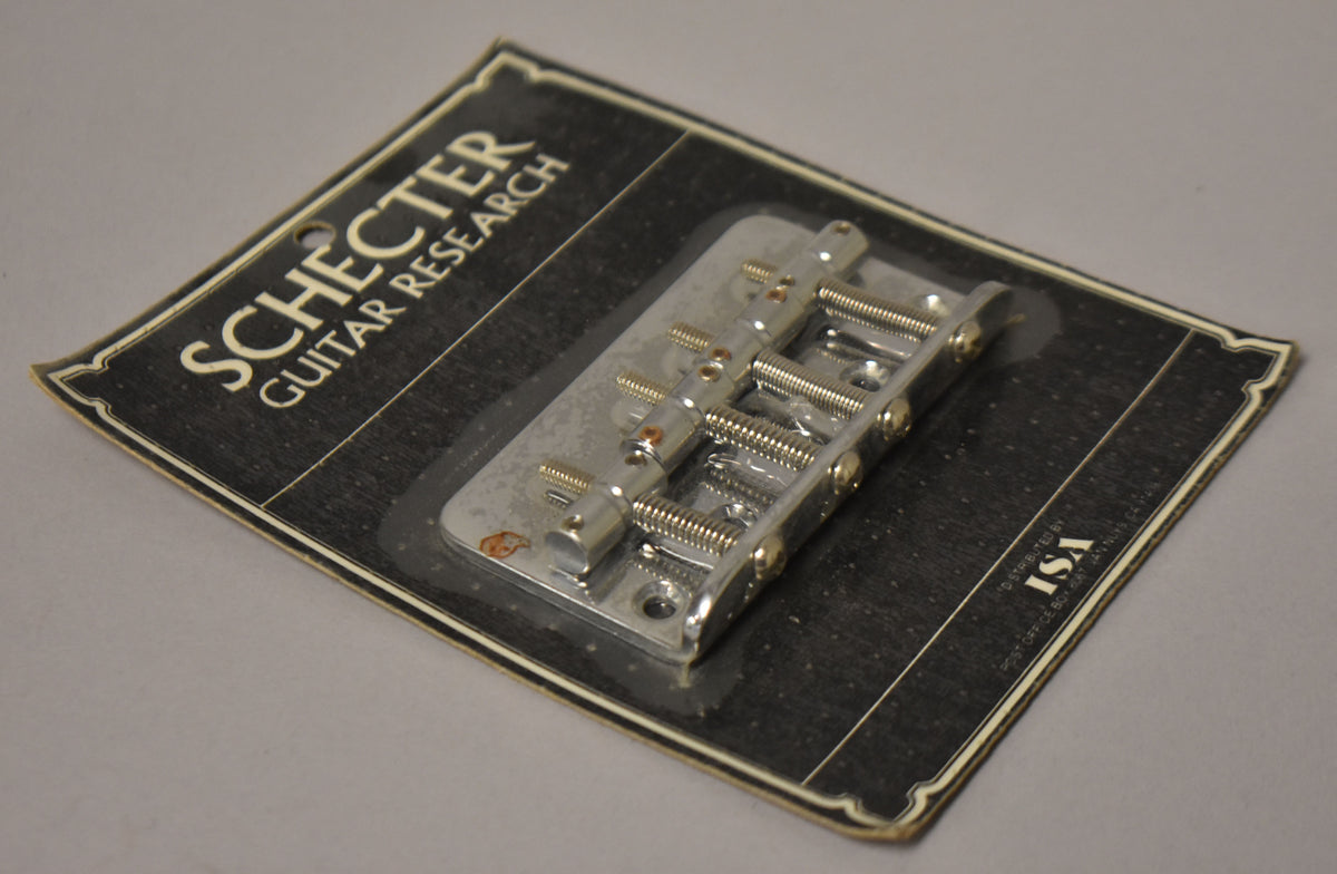 A chrome 4-string bass bridge in blister packaging, angled from the top right, with visible rust spots on the baseplate.
