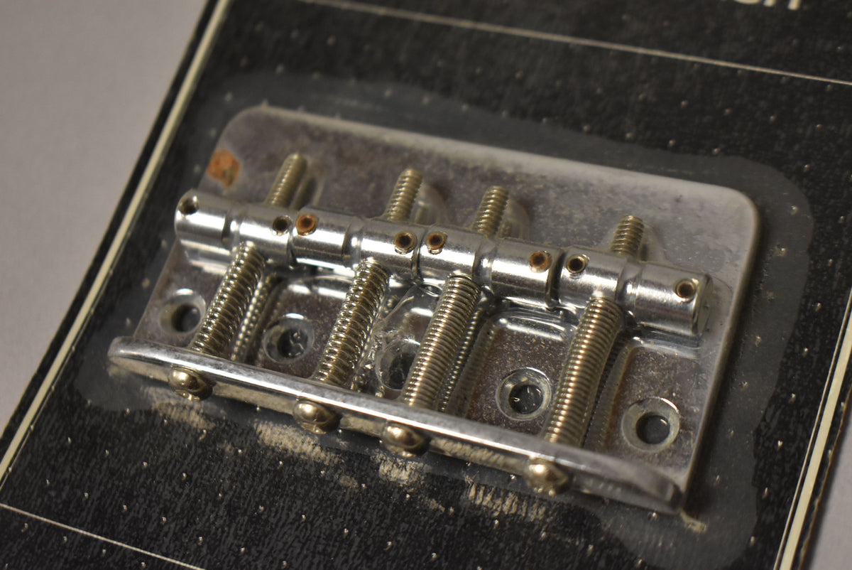 Close-up, top-down view of a chrome 4-string bass bridge with four adjustable saddles, showing some tarnishing on the screws.