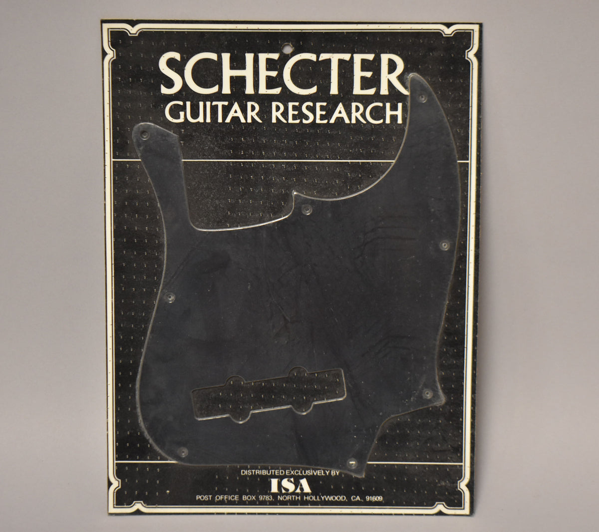 Black anodized J-style bass pickguard, still sealed in its original clear plastic packaging, shown from a slightly angled front view.