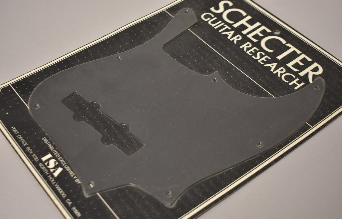 A black anodized J-style bass pickguard with a matte finish, showing some light surface scuffs, still sealed in its original packaging.