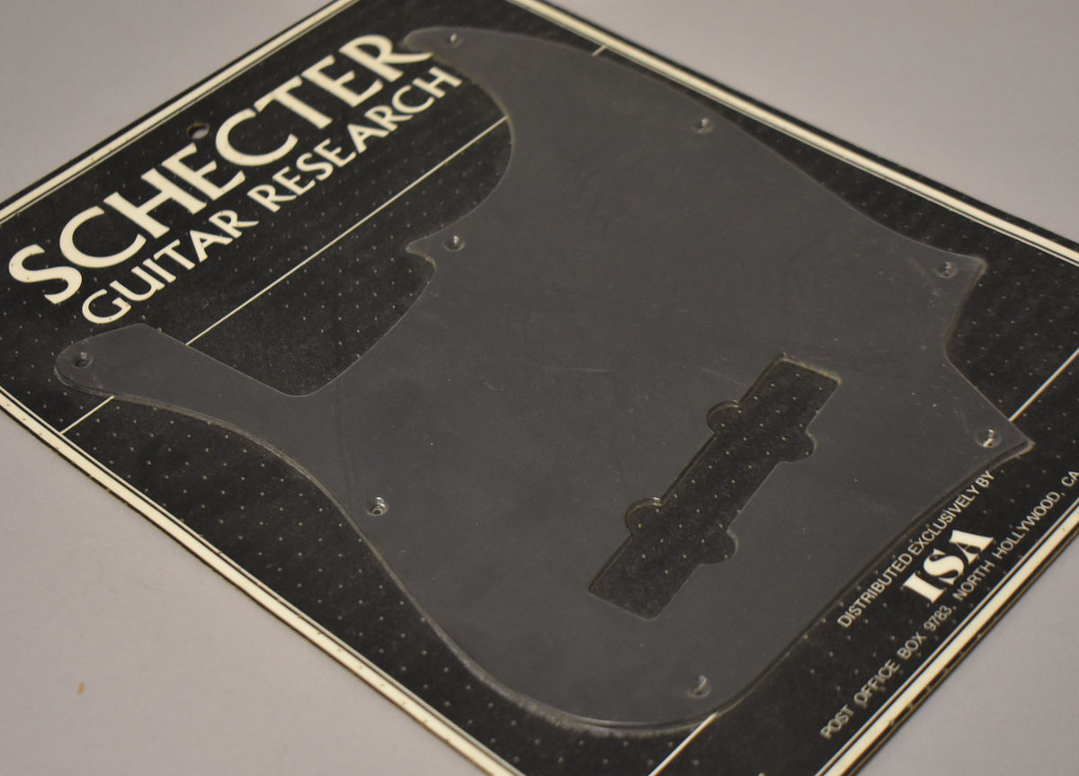 A black anodized J-style bass pickguard with a P-bass pickup route, still sealed in its original clear plastic packaging.