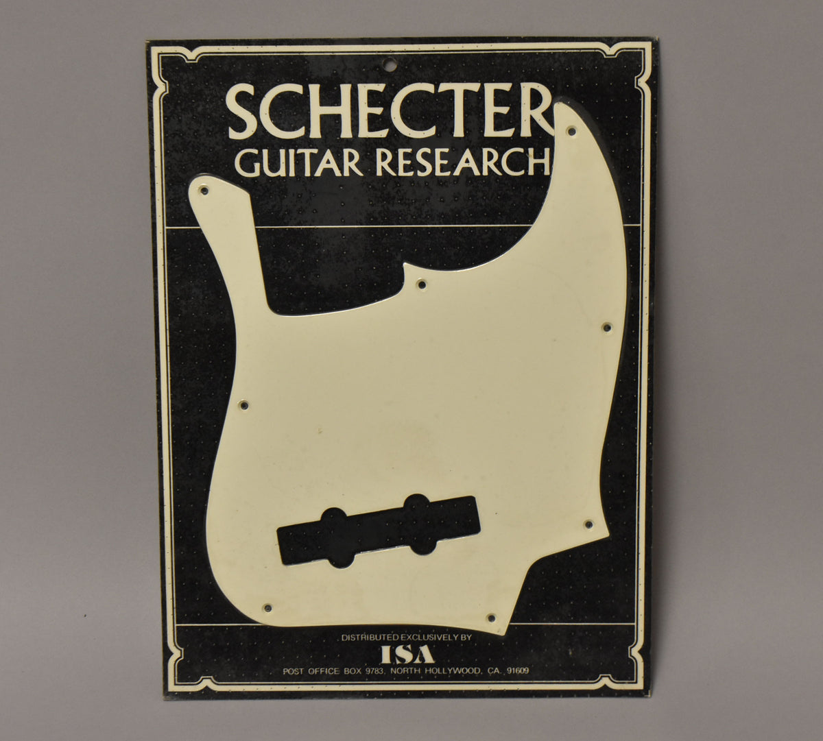 A full-front view of a white J-style bass pickguard with a sealed surface, mounted on a black speckled display card.