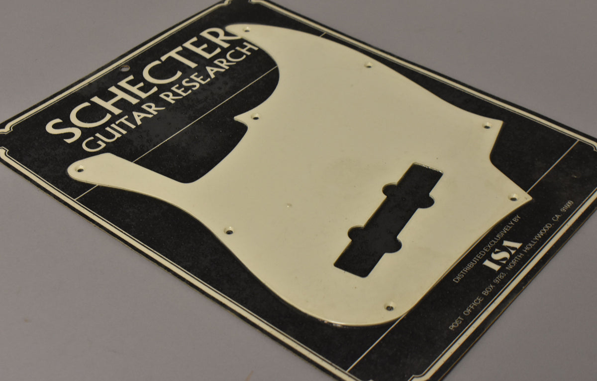 The off-white J-style bass pickguard, still sealed in its original black and cream packaging, is shown from a high-angle view.