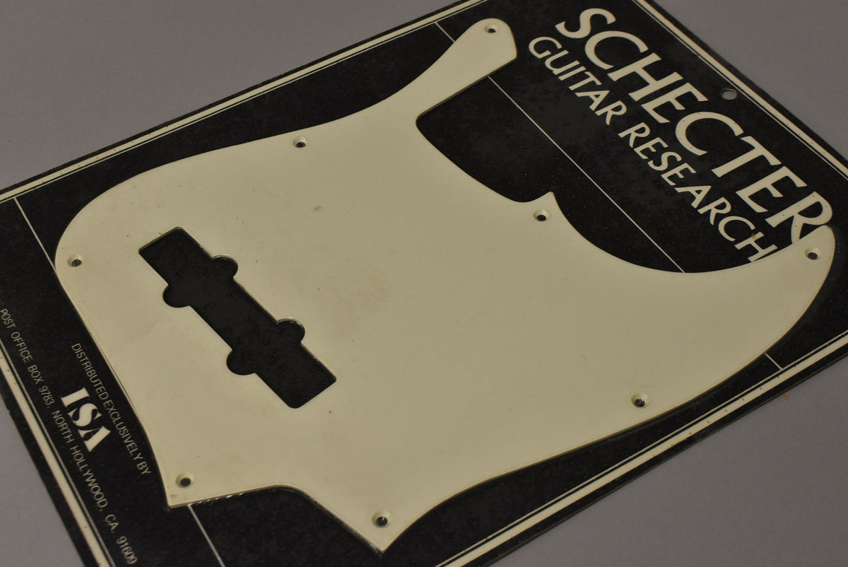 The off-white pickguard, with a P-bass pickup cutout, is sealed in black packaging and viewed from a high angle.