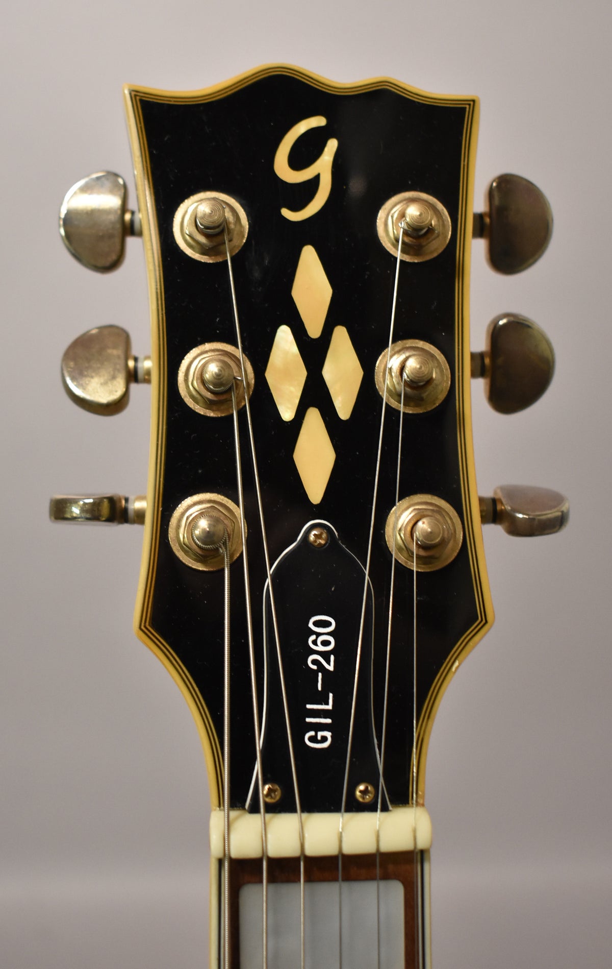 Close-up of the black headstock with gold "G" logo, diamond inlays, six gold tuners, and "GIL-260" on the truss rod cover.