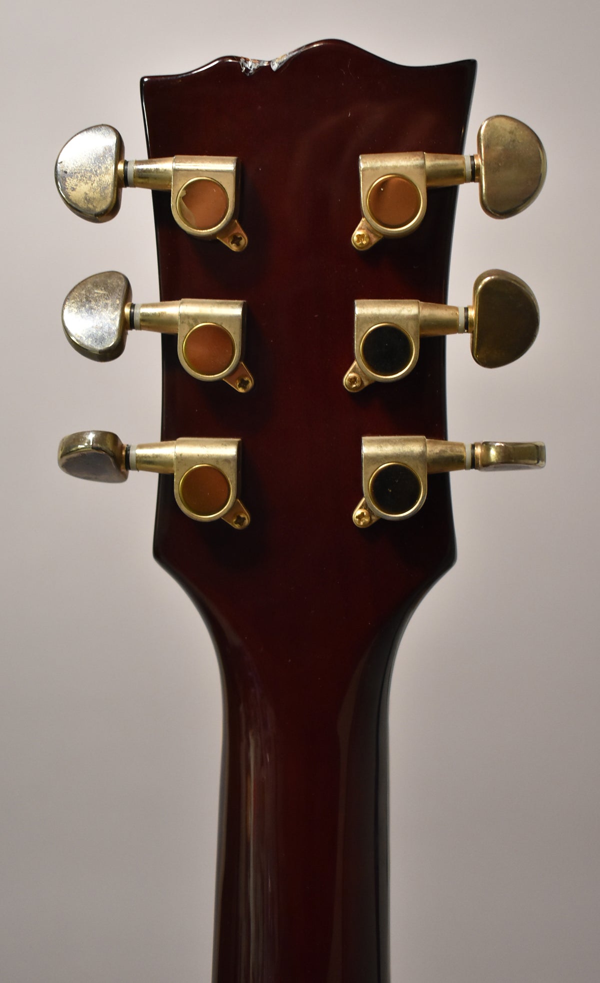 The back of a dark brown headstock with six gold-plated tuners, showing wear and chips along the top edge.