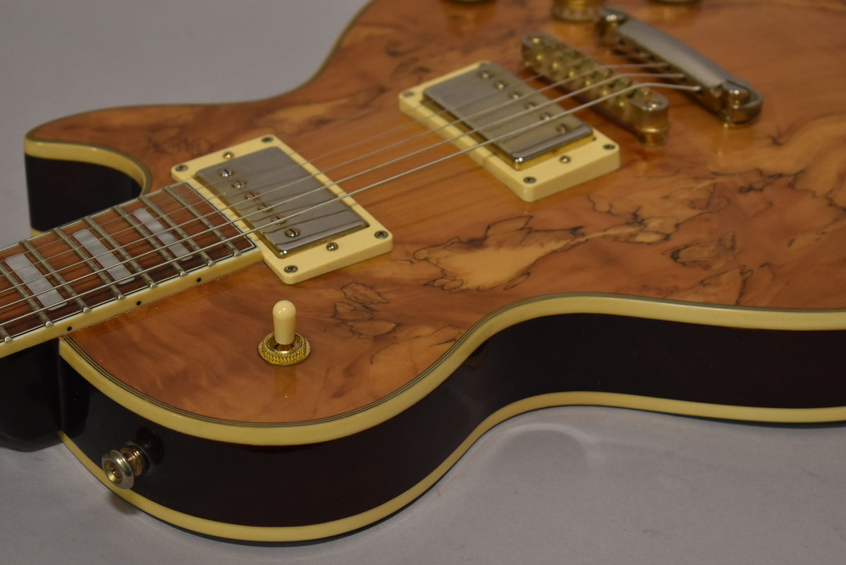 Close-up of the guitar's body, showing the natural burl wood finish, cream binding, two humbuckers, and a cream toggle switch.