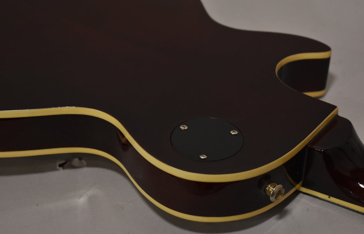 The lower bout of a dark brown guitar, showing the back control cavity cover, output jack, and cream binding.