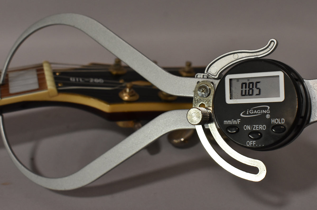 A digital caliper measures the nut width of the guitar's dark brown headstock, showing "0.85" on its screen.