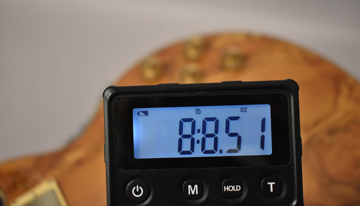 A close-up of a digital scale displaying "8:8.51 lb oz" in blue, with the guitar's natural finish body blurred in the background.