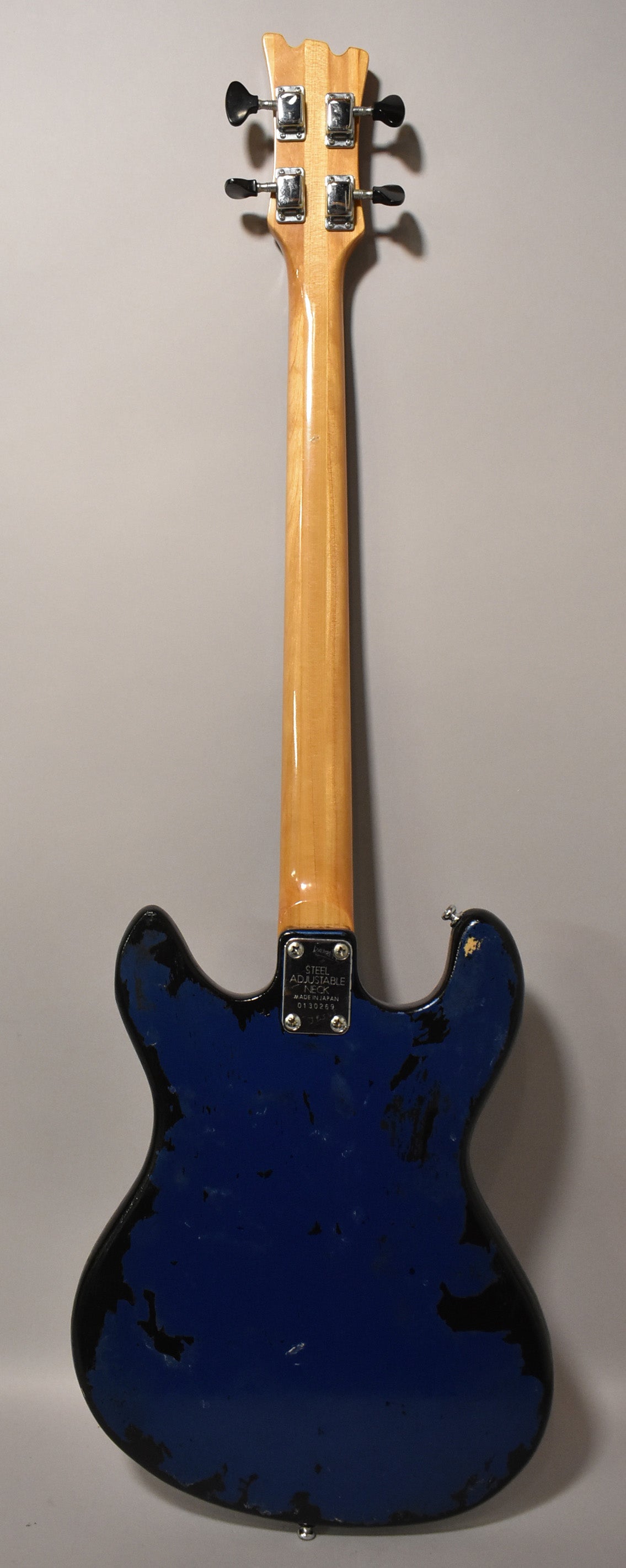 Rear view of a metallic blue bass with heavy paint chipping, a light wood neck, and four chrome tuners with black knobs.