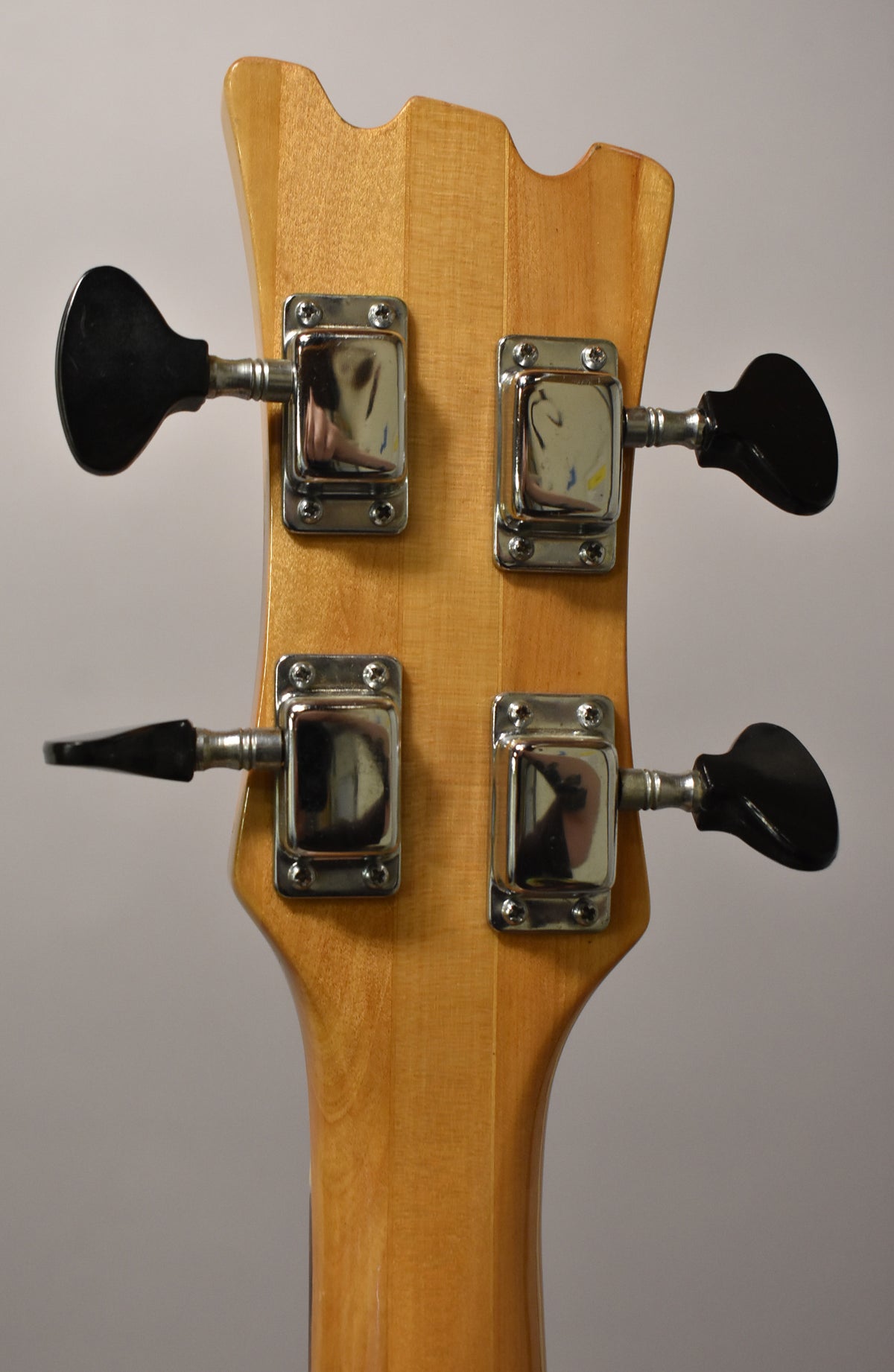 Rear view of the bass headstock and top of the neck, showing four chrome tuners with black paddle keys on a light wood finish.