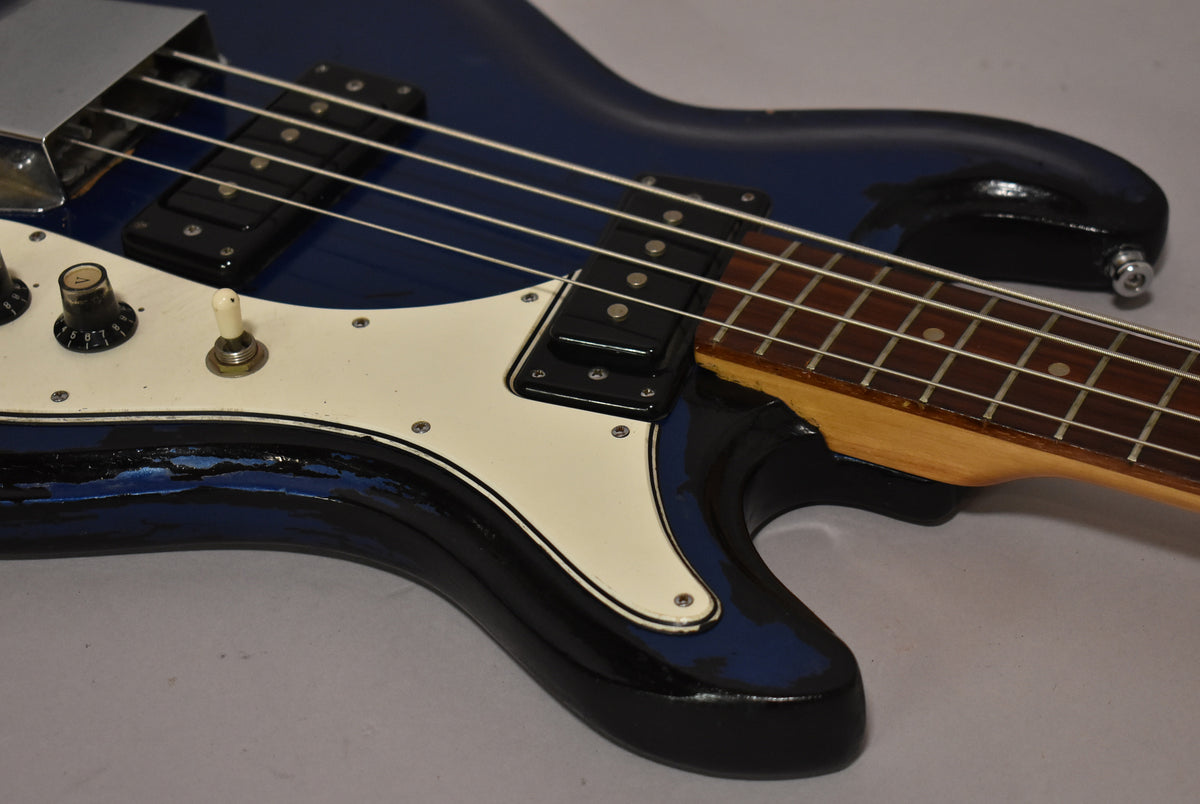 Close-up of the bass body, showing the metallic blue finish with heavy wear, white pickguard, two black knobs, and a white switch.