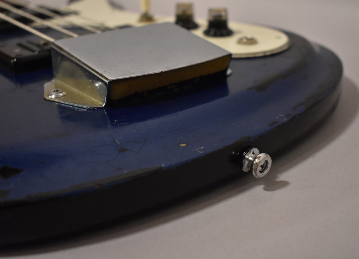 Close-up of the bass body, showing the metallic blue finish with heavy wear, a chrome bridge cover with foam, and a strap button.