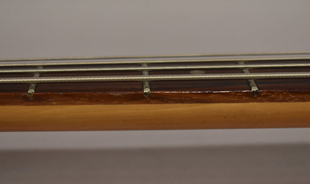 Close-up of the bass neck, showing four strings over a dark fretboard with light fret markers and a worn, light-colored neck.