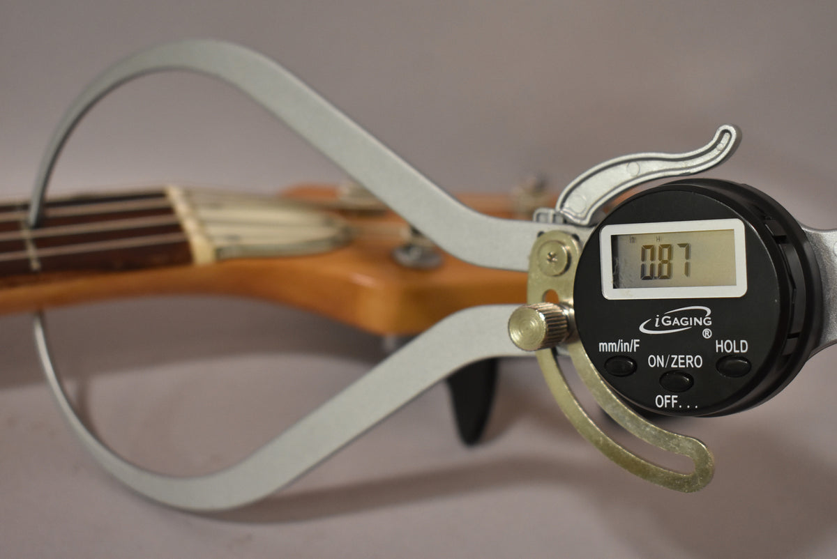 A digital caliper measures the bass's neck width at the nut, displaying "0.87" on its screen.