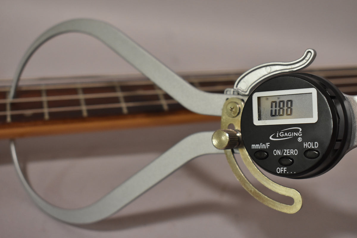 A digital caliper measures the bass's fretboard and frets, displaying "0.00" on its screen.