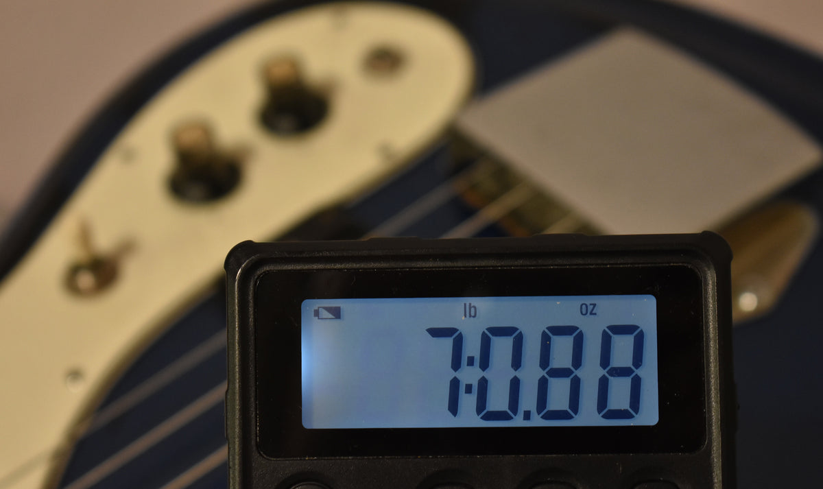 A digital scale displays "7 lb 0.88 oz" in the foreground, with a blue bass body and cream pickguard blurred behind it.