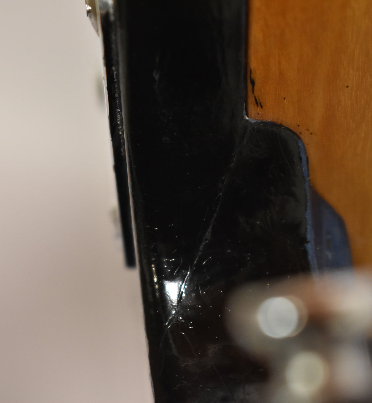 Close-up of the bass's black headstock, showing finish checking and a chip near the nut, with a blurred tuning peg in the foreground.