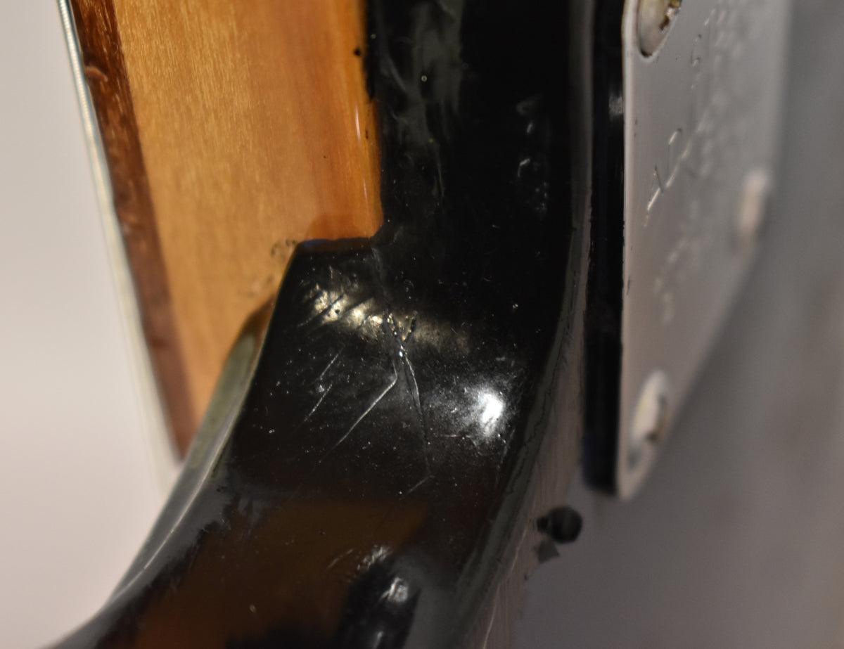 Close-up of a guitar neck joint, showing a black finish with scratches and a light wood body, next to a metal plate.