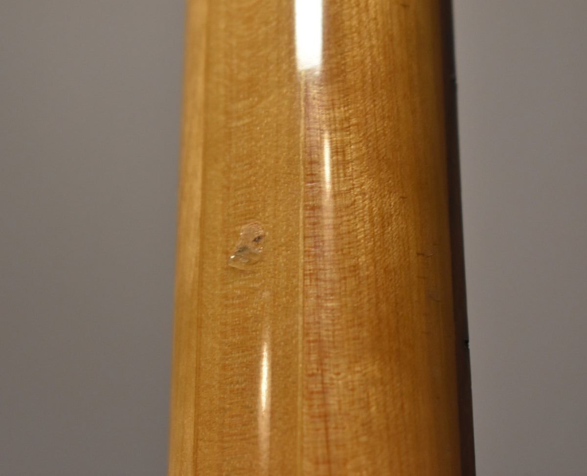 Close-up of the bass neck's light brown wood grain, showing a small, clear chip in the glossy finish near the center.