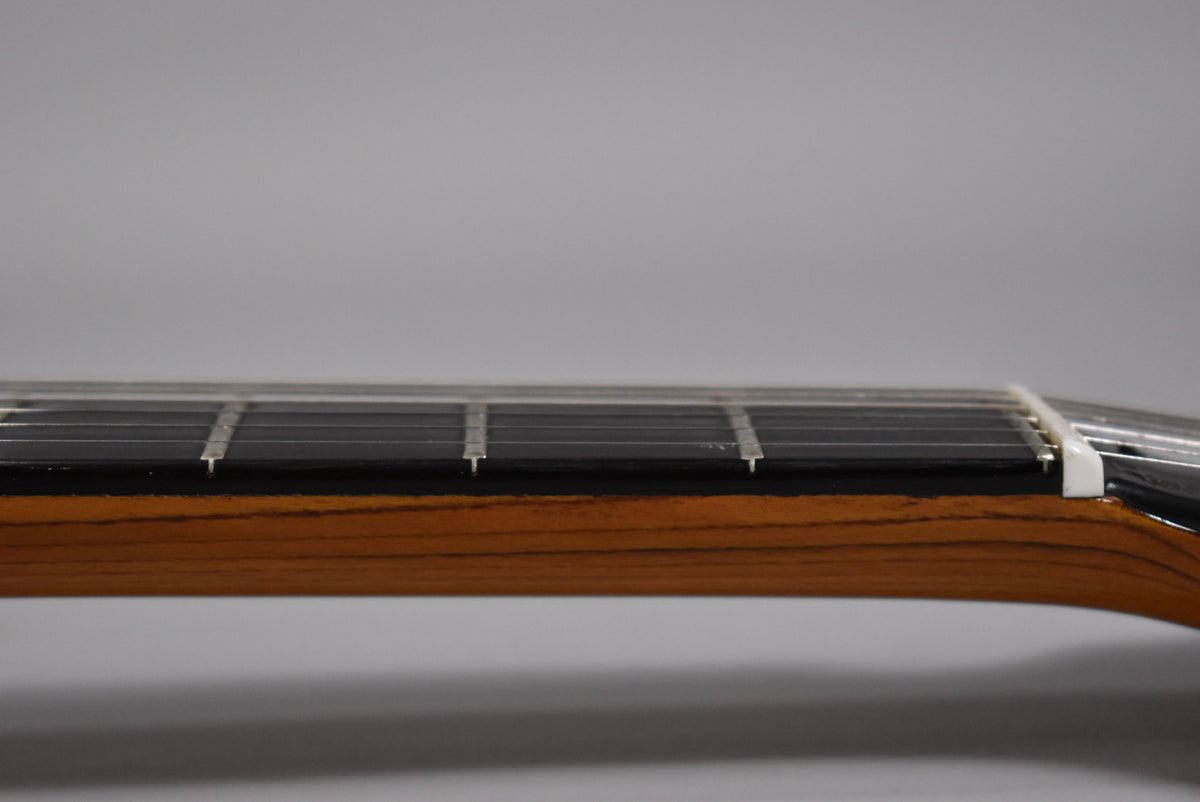 Side view of the guitar neck, showing the dark fretboard, silver frets, and a white nut, with a rich brown wood grain.