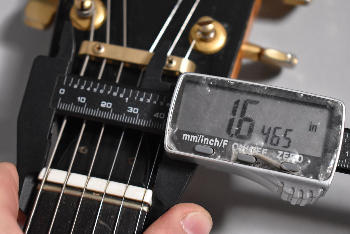 A close-up of the guitar's nut and first fret, with a digital caliper measuring the nut width at 1.6465 inches.