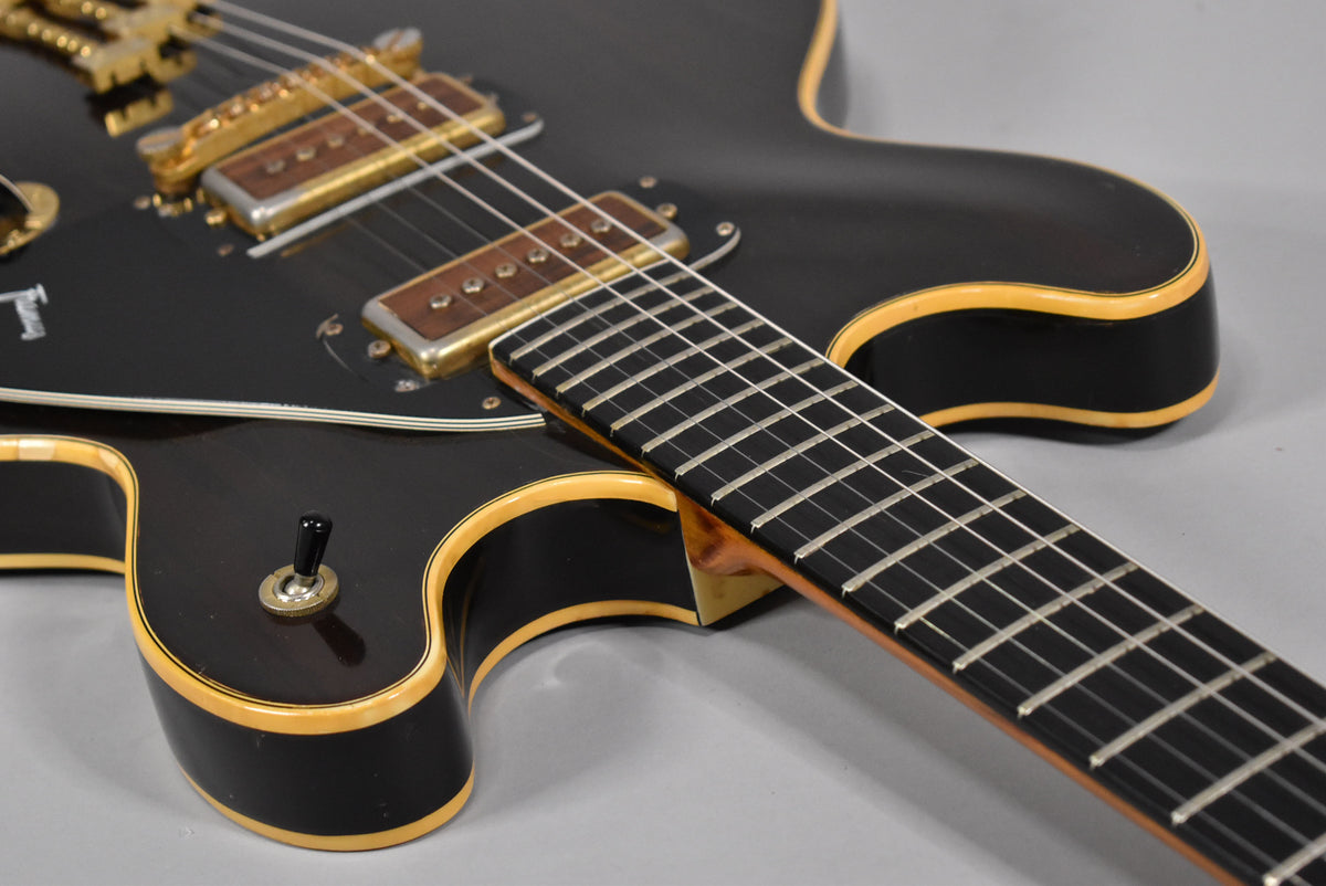 Close-up of a dark walnut finish hollow body electric guitar, showing the lower bout, black pickguard, gold pickups, and fretboard.