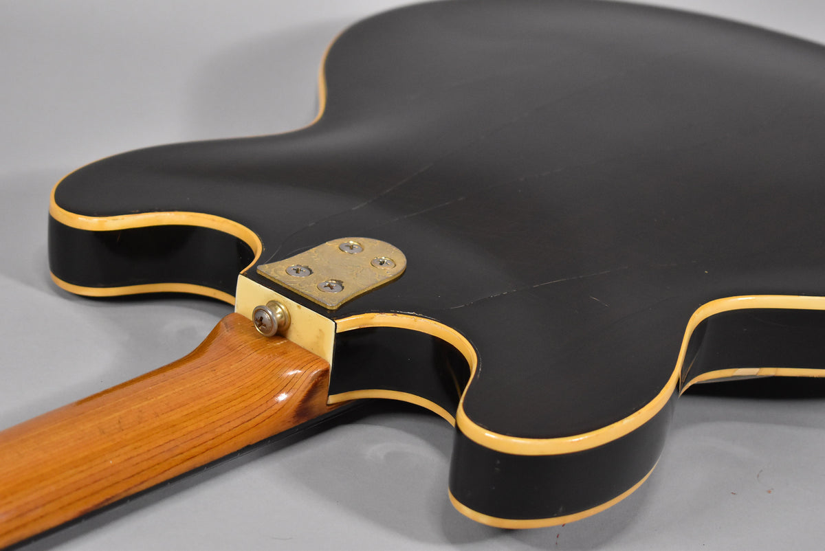 The guitar's black body, with cream binding and visible finish checking, meets a light wood neck with a brass neck plate.