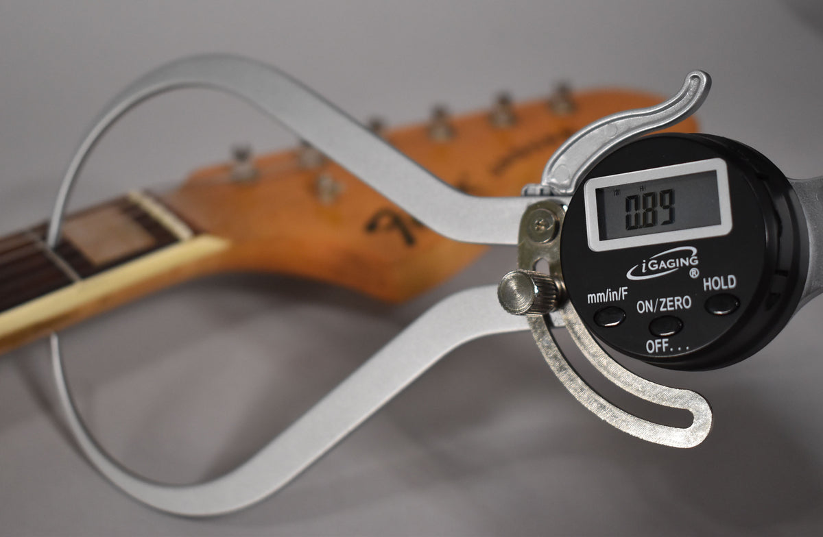A digital caliper measures the neck thickness at the first fret, showing 0.89 inches, with the headstock blurred in the background.