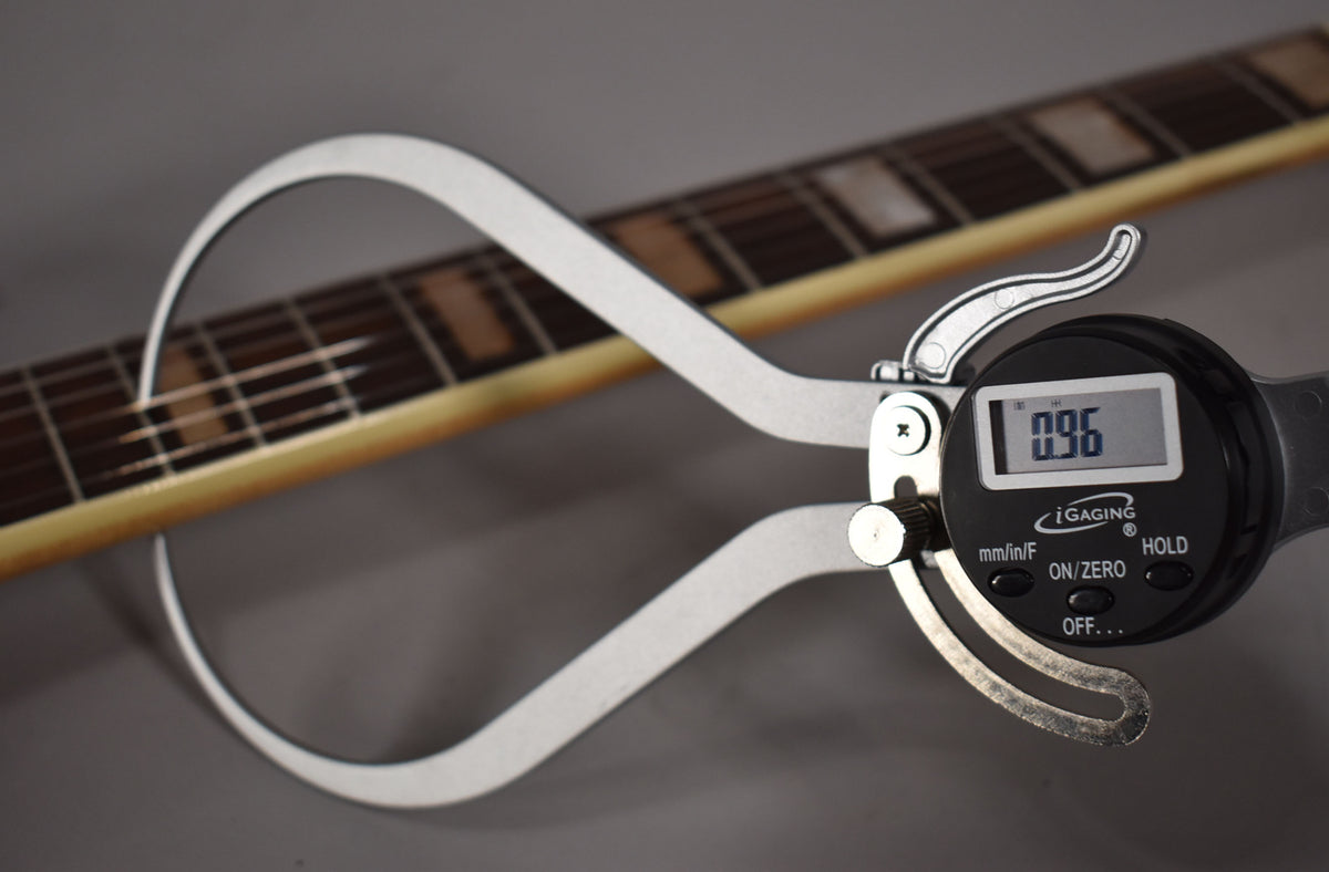 A digital caliper measures the guitar's fretboard, showing 0.96, with block inlays visible on the dark wood fretboard.