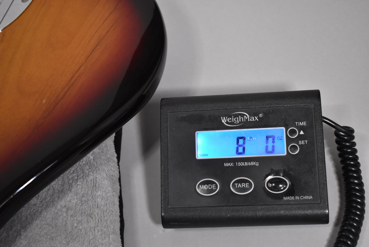 The guitar's sunburst body, showing some light scratches, rests on a grey towel next to a digital scale displaying "8 lb 0 oz".