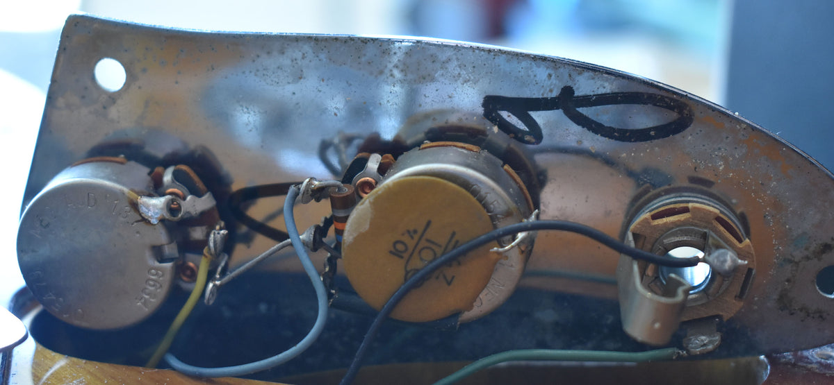 Close-up of the guitar's control cavity, showing three tarnished potentiometers, a yellow capacitor, and wiring on a worn metal plate.