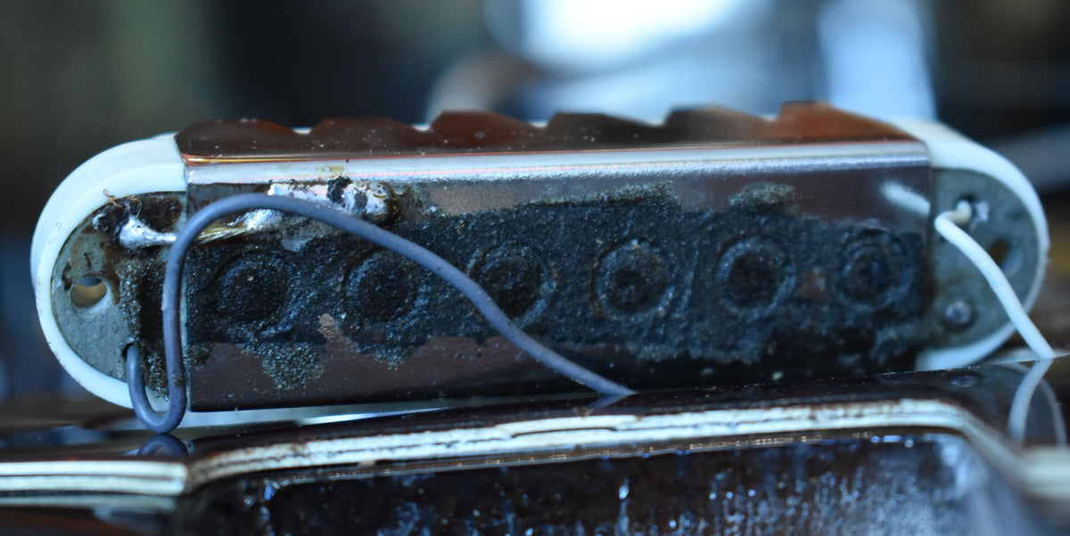 Close-up of a chrome-covered pickup with exposed black wax potting, white bobbin ends, and visible wiring, showing heavy wear and grime.