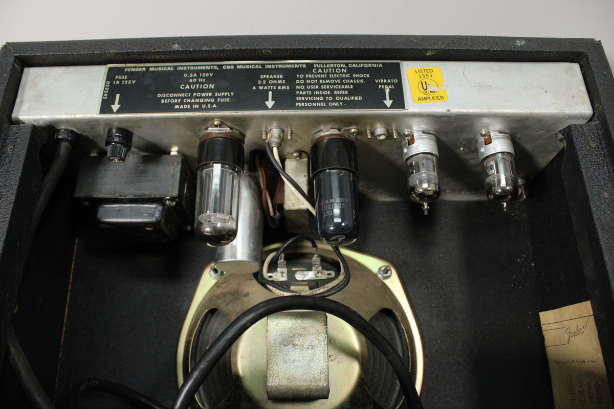 Rear view of the amp's chassis, showing four glowing vacuum tubes, a silver control panel, and the top of a gold-framed speaker.