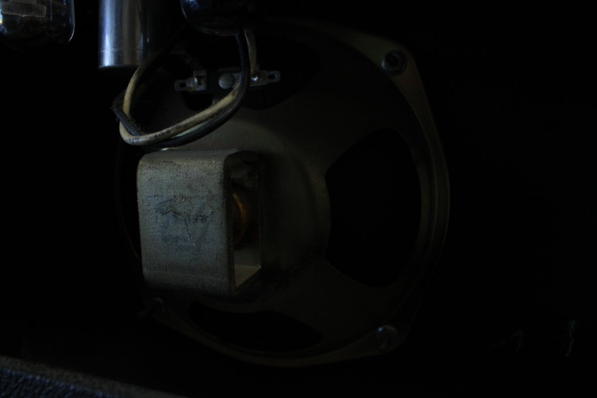 A dark, close-up view of the amp's internal speaker and wiring, showing the speaker's metal frame and magnet.