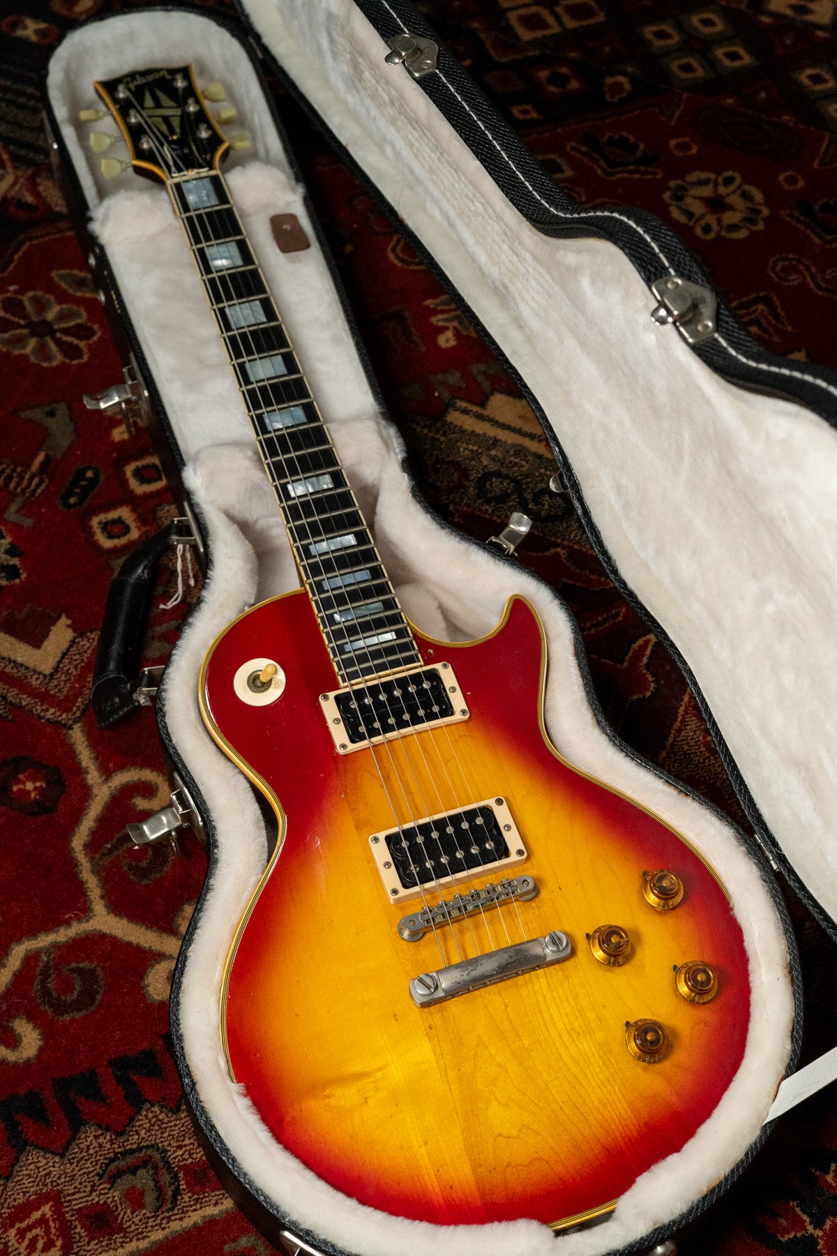 A cherry sunburst Les Paul Custom in a white fur-lined hard case, showing heavy checking and wear on its body and gold hardware.