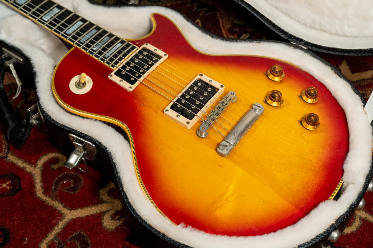 Cherry sunburst guitar body, angled in its white-lined case, shows two humbuckers, four amber knobs, and a worn bridge.