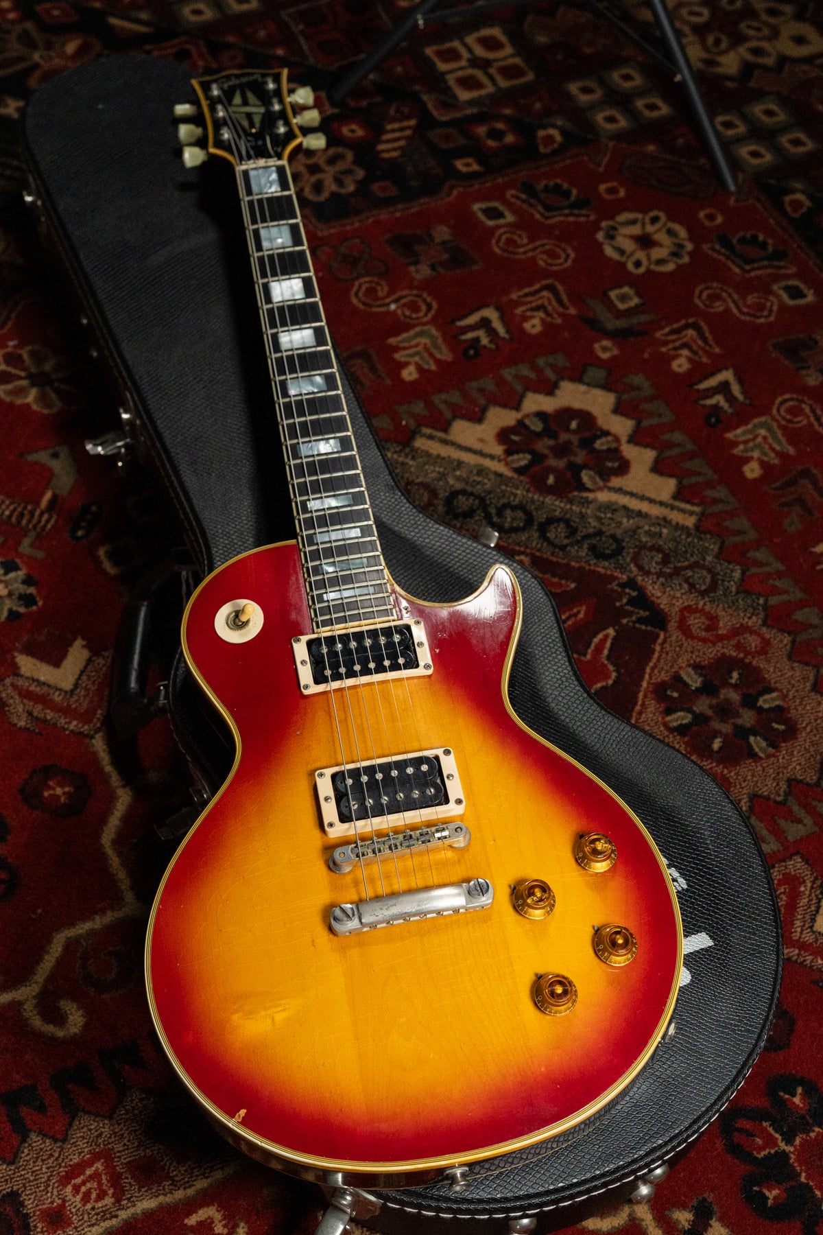 Full-length, top-down view of a cherry sunburst electric guitar with cream binding, gold hardware, and block inlays, resting on its open hard case.