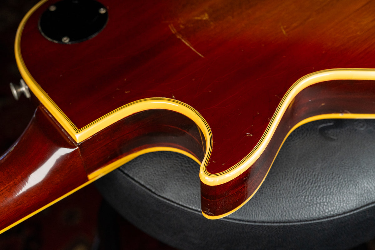 Close-up of the guitar's lower bout and neck heel, showing a cherry sunburst finish with binding, scratches, and a strap button.