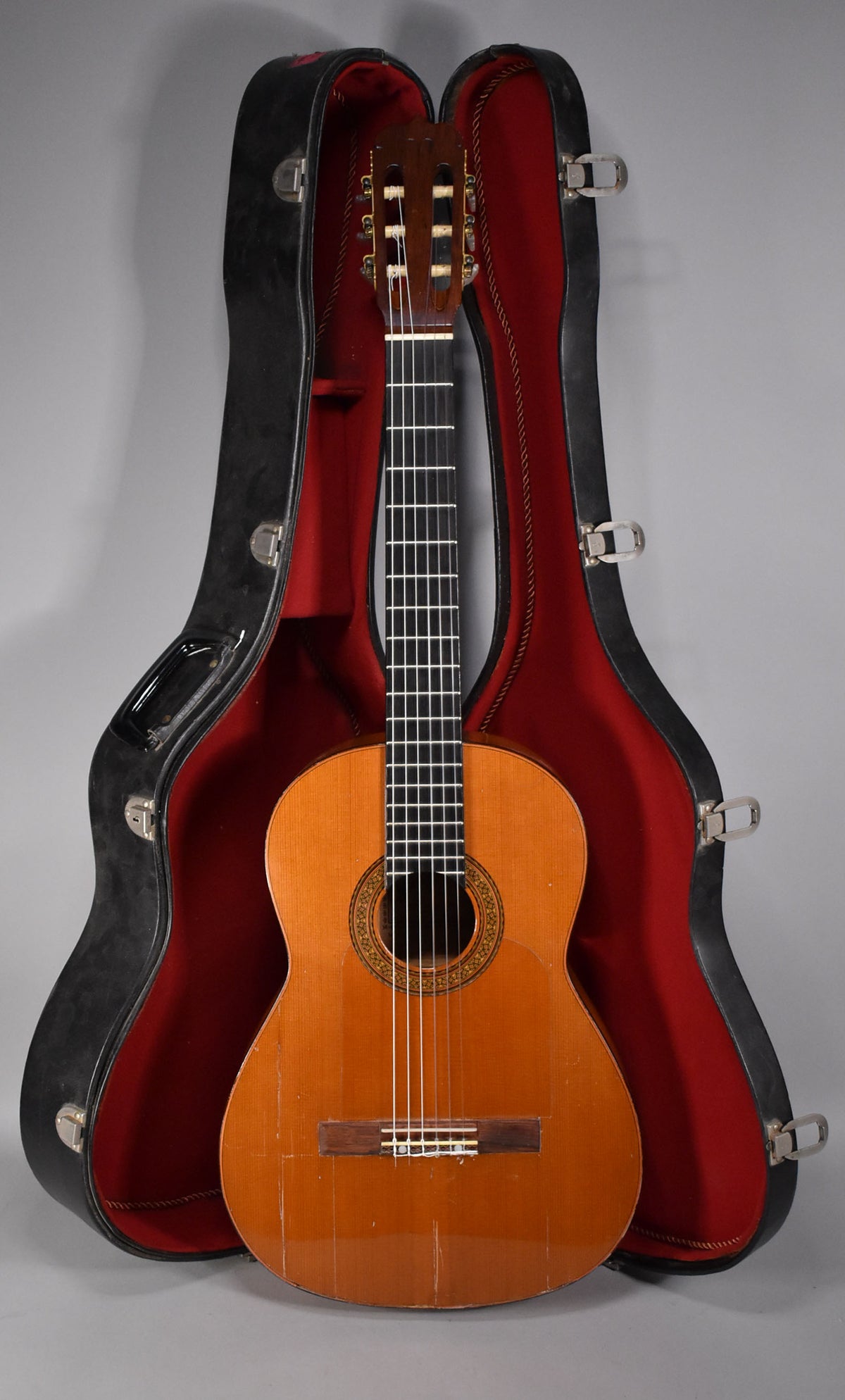 Full-length view of a natural finish classical guitar with a dark fretboard, standing in its open black hard case with red lining.