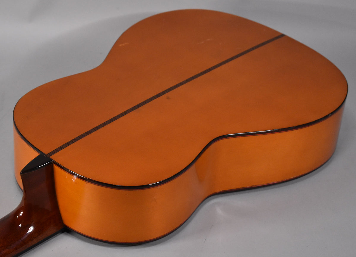 The classical guitar's natural finish back and sides are visible from the lower bout, showing a dark center strip and some light wear.