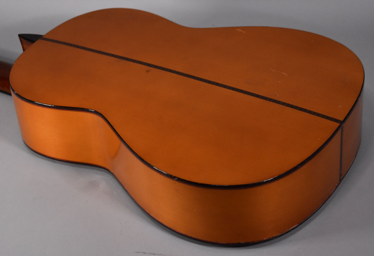 The classical guitar's natural finish back and sides are visible, showing a dark center strip and binding, with light wear and scratches.