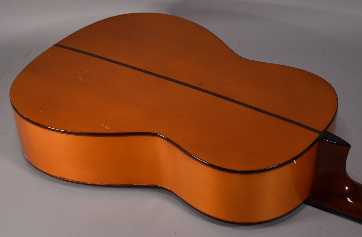 The classical guitar's natural finish back and side are visible from an angle, showing a dark center strip and some minor scratches.