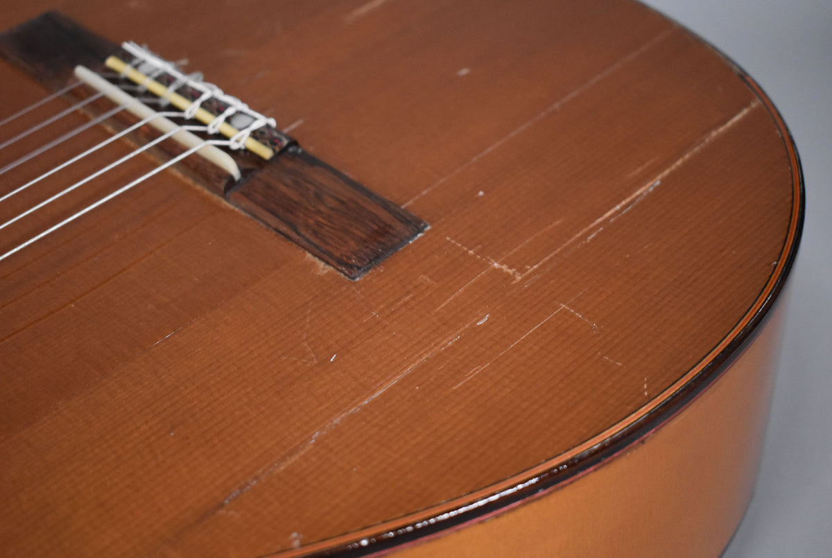 Close-up of the classical guitar's natural finish top, showing a dark wood bridge, white saddle, and numerous scratches and dings.